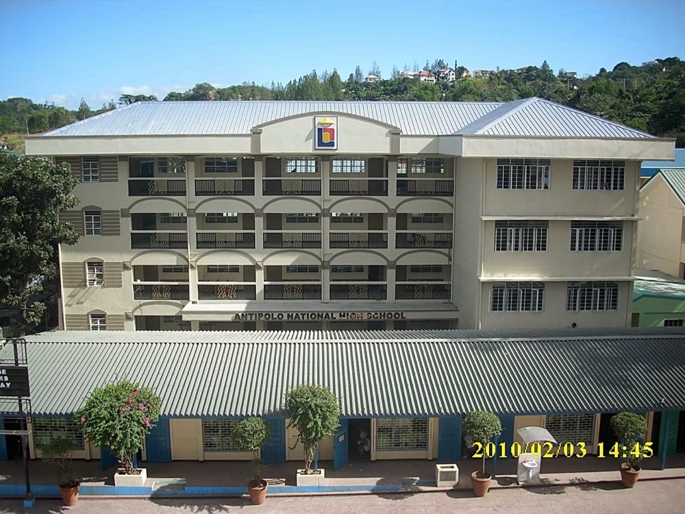 Completed multi-storey school building: long rows of windows and open circulation corridors across a pale rendered facade.
